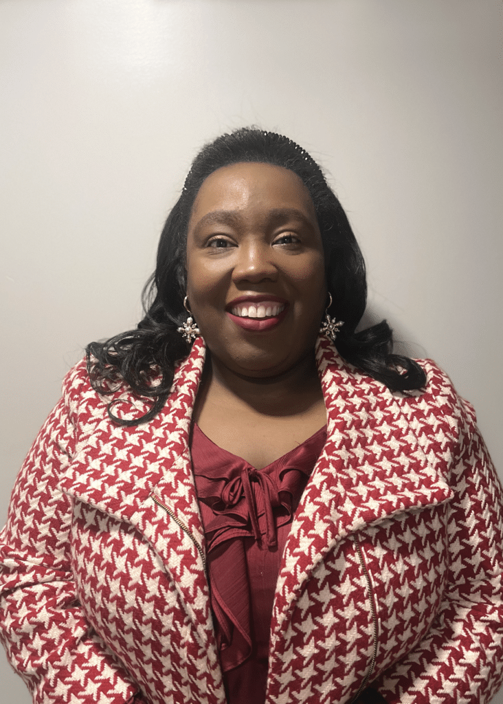 Woman smiling, wearing a red and white houndstooth jacket and maroon top, standing against a plain background.