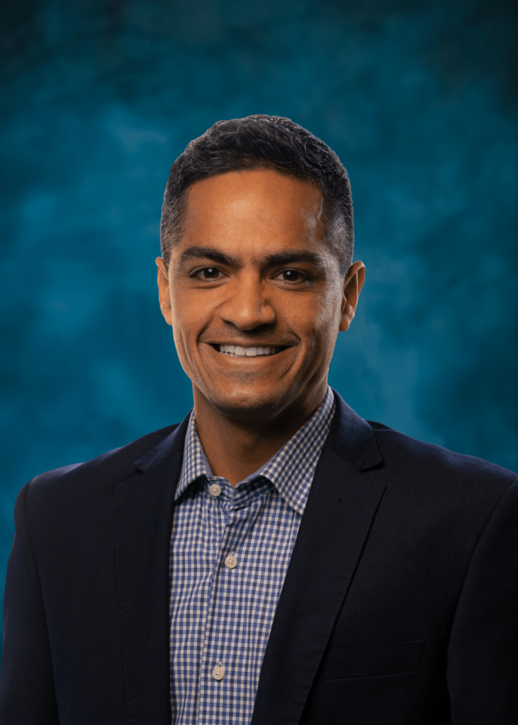Smiling man in a suit jacket and checkered shirt, posed against a blue background.
