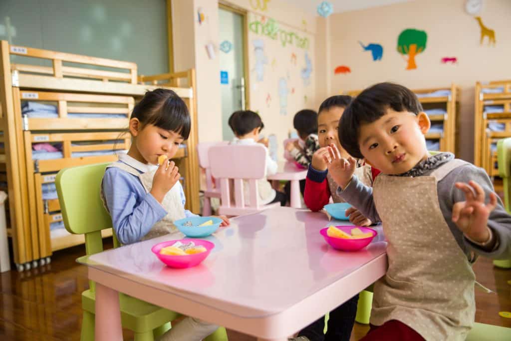 Three young children sit at a pink table in a classroom, eating snacks from colorful bowls.