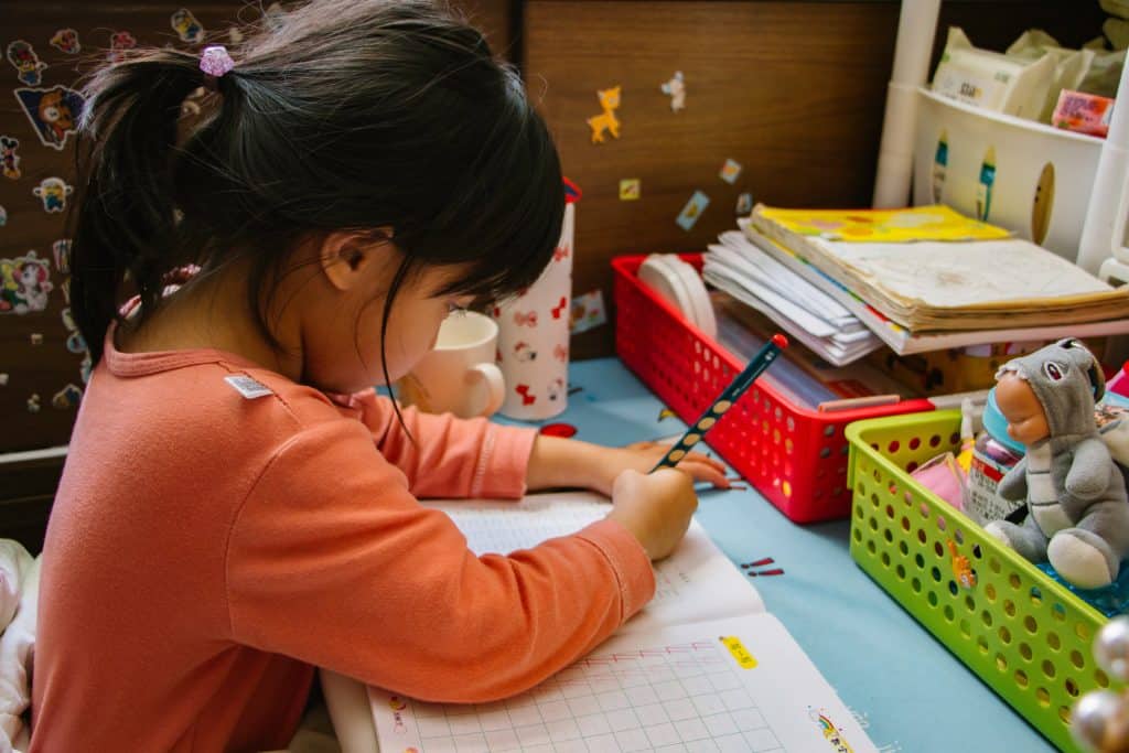 Young girl with dark hair writing in a notebook at a desk covered with books, papers, and colorful baskets.