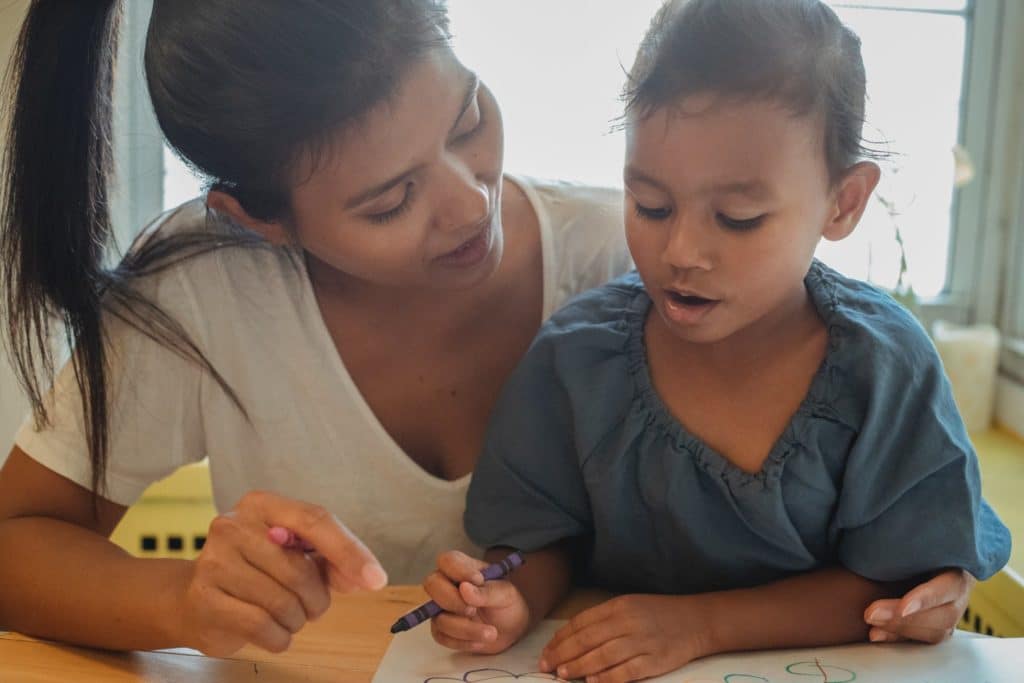 Woman helping young girl color with crayons at a table, both focused on the drawing.