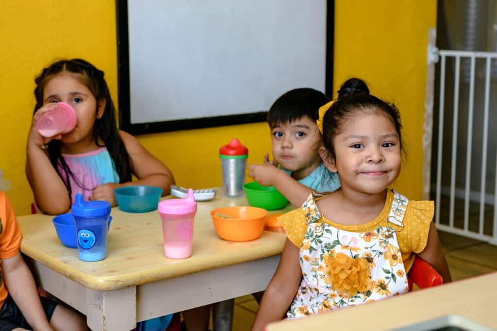 Three young children sit at a small table, eating snacks and drinking from cups, with a yellow wall behind them.