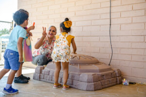Woman smiling and flashing a peace sign as two children play with chalk near a sandbox outside.