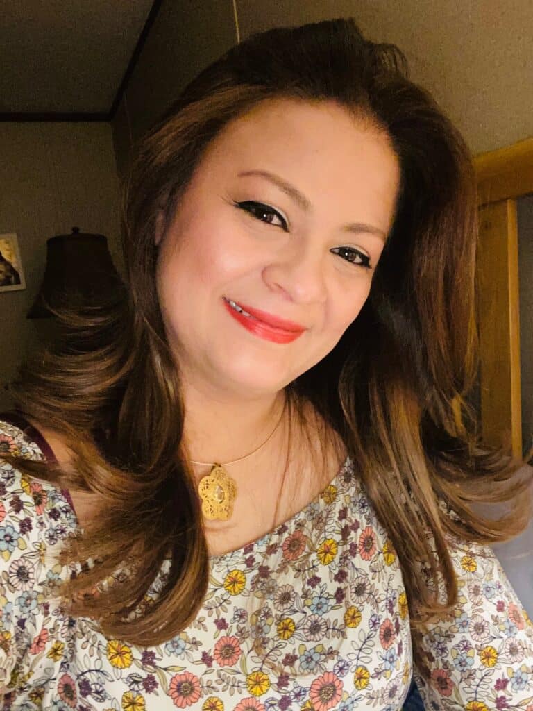 Smiling woman with long brown hair, wearing a floral top and gold necklace, sitting indoors.