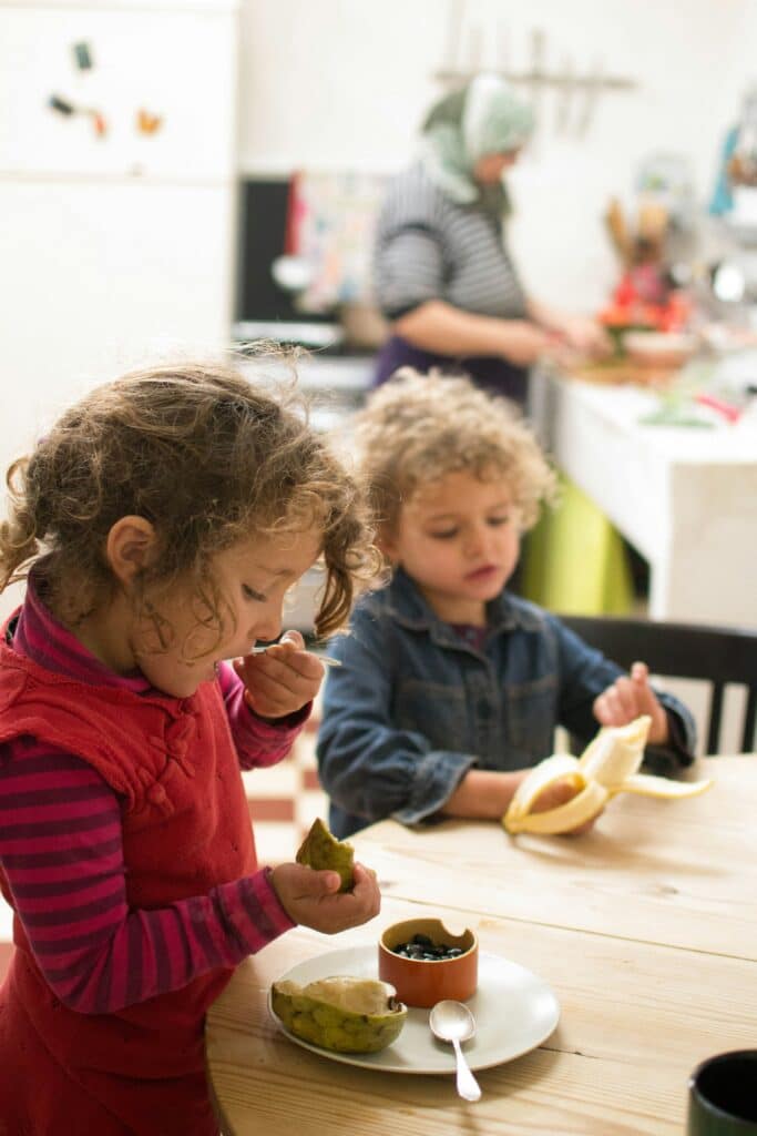 Two young children eat fruit at a table while an adult prepares food in the background kitchen.