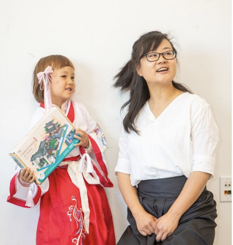 A child in a costume holds a toy box, smiling next to a woman in glasses and a white shirt.