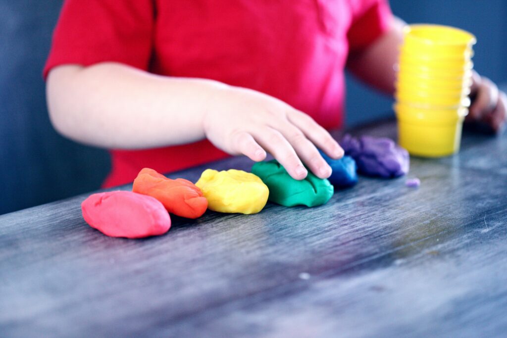 A child in a red shirt touches colorful playdough arranged in a row on a table.