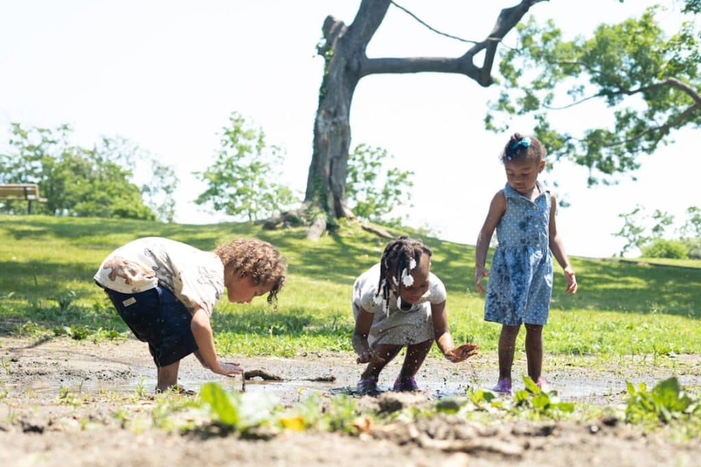 Three young children play and explore in a muddy area in a sunny park with green grass and trees.