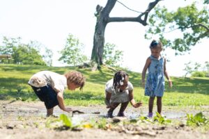 Three young children play and explore in a muddy area in a sunny park with green grass and trees.