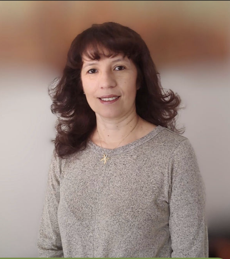 Woman with wavy brown hair wearing a grey sweater, smiling, standing in front of a blurred background.