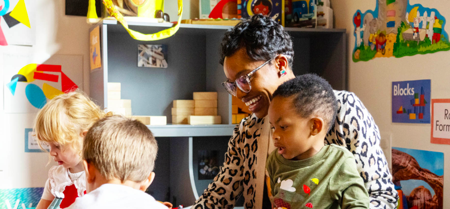 An adult and three young children play together at a table in a colorful classroom setting.