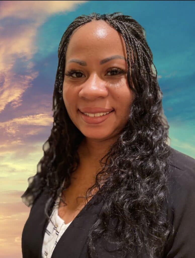 Smiling woman with long braided hair, wearing a dark blazer, stands in front of a colorful sunset sky.