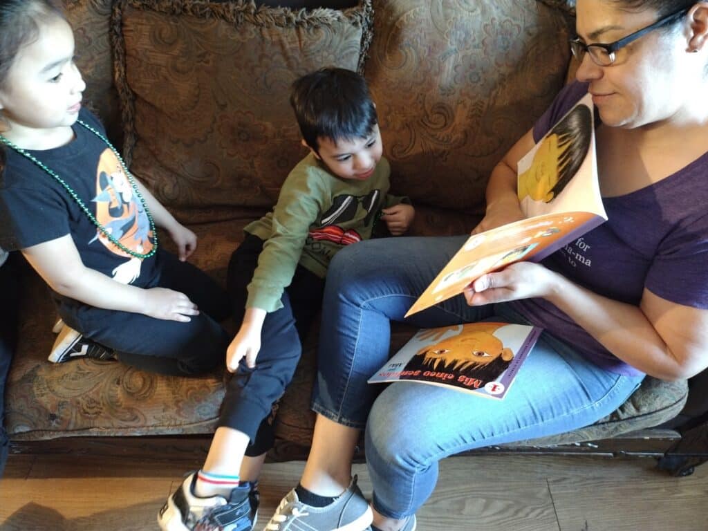 A woman reads a book to two children sitting with her on a patterned couch.