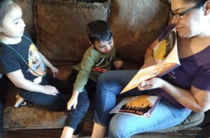 A woman reads a book to two children sitting with her on a patterned couch.