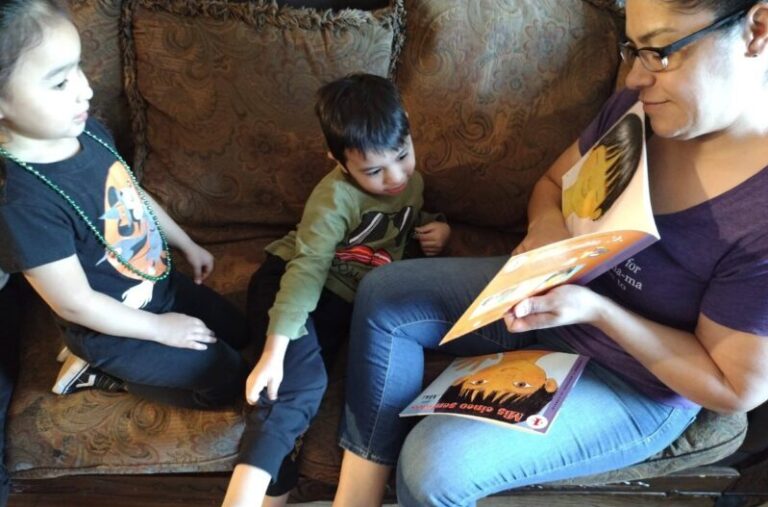 A woman reads a book to two children sitting with her on a patterned couch.