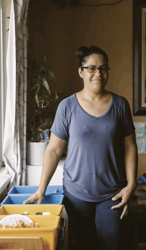 Woman in glasses and a blue shirt stands indoors by colorful bins near a window with a houseplant.