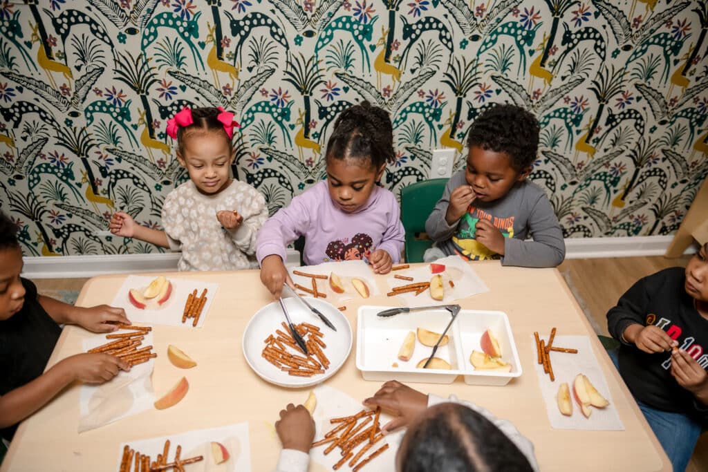 Five young children sit at a table, using tongs to pick up pretzels and apple slices from plates.