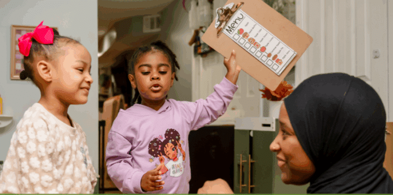 Two young girls talk to a woman in a hijab; one girl holds up a clipboard with a menu chart.