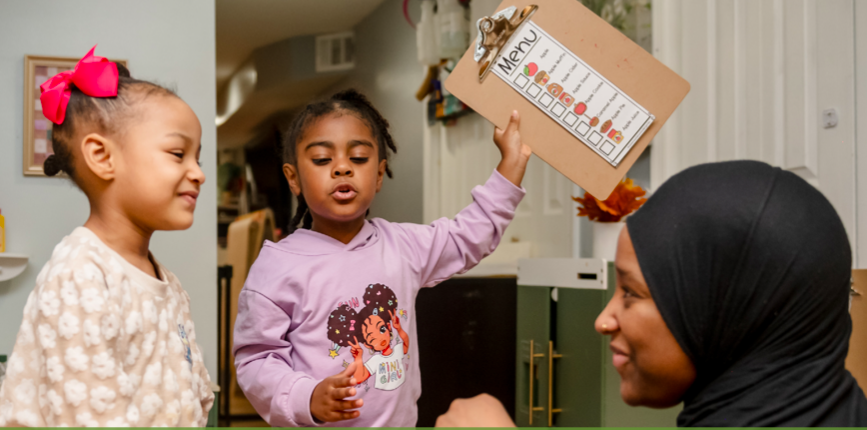 Two young girls talk to a woman in a hijab; one girl holds up a clipboard with a menu chart.