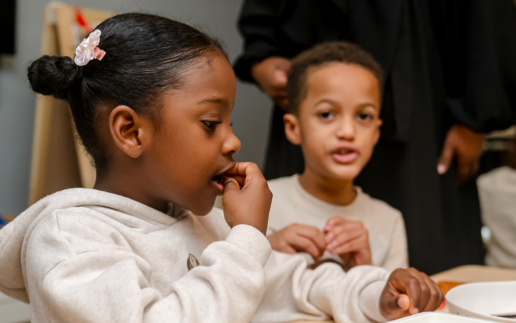 Two young children sit at a table, one eating and the other talking, both wearing light-colored hoodies.