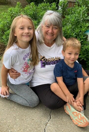 Smiling older woman sitting outside with two young children on a sidewalk, greenery in the background.