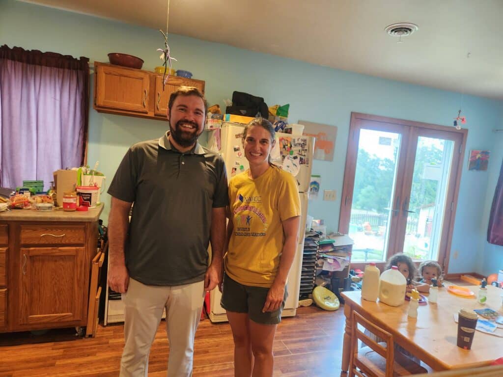 Two adults smile in a kitchen while children sit at a table in the background near a glass door.