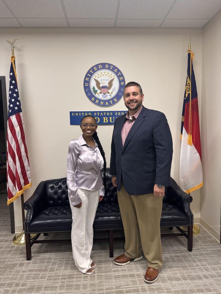 Two people smiling and standing in front of a couch with U.S. Senate and state flags in an office.