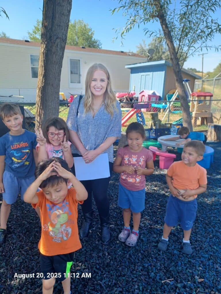 A smiling woman stands outdoors with six happy children at a playground on a sunny day.