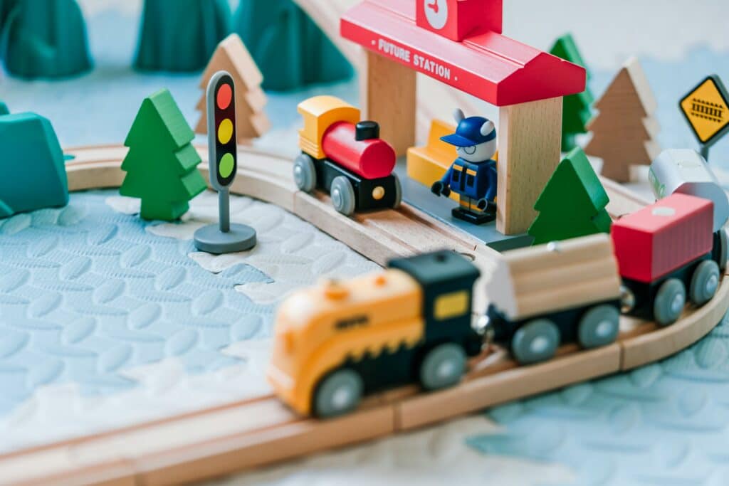 Wooden toy trains on tracks near a station, traffic light, trees, and a toy figure in a blue uniform.