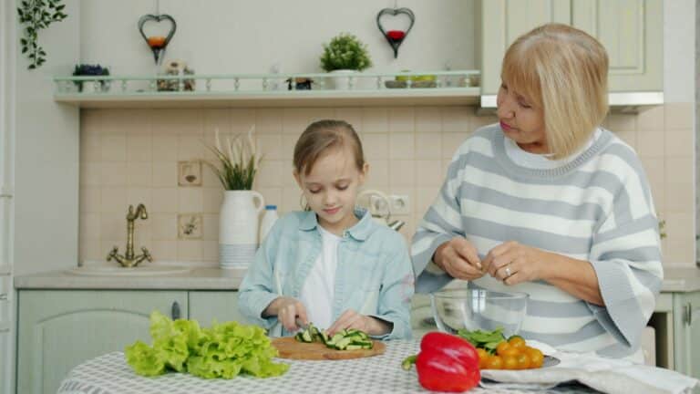 A young girl slices cucumbers while an older woman prepares salad in a kitchen with fresh vegetables.