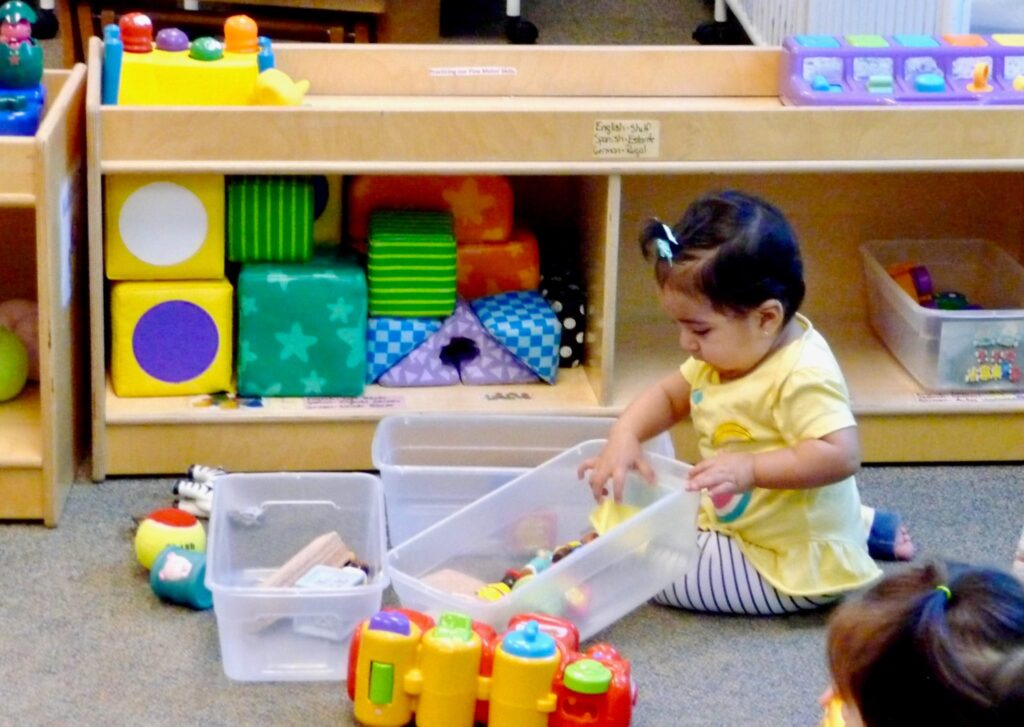 A toddler in a yellow shirt sits on the floor, playing with toys and bins in a colorful playroom.