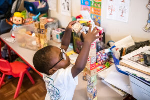 A young child stacks wooden blocks on a tall tower in a colorful, cluttered playroom.