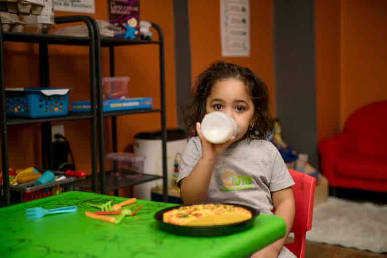 A young child sits at a green table, drinking from a bottle, with a plate of food in front of them.