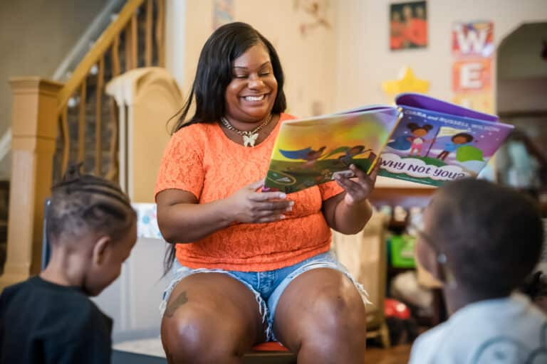 Woman reading a colorful book to two young children in a cozy, home-like setting.