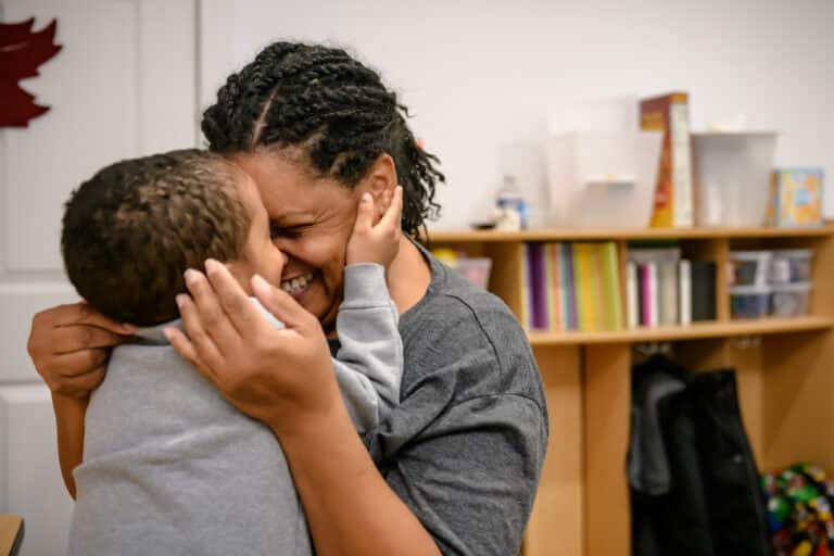 Adult and child smiling and hugging closely in a warm, joyful moment in a cozy room with bookshelves behind them.