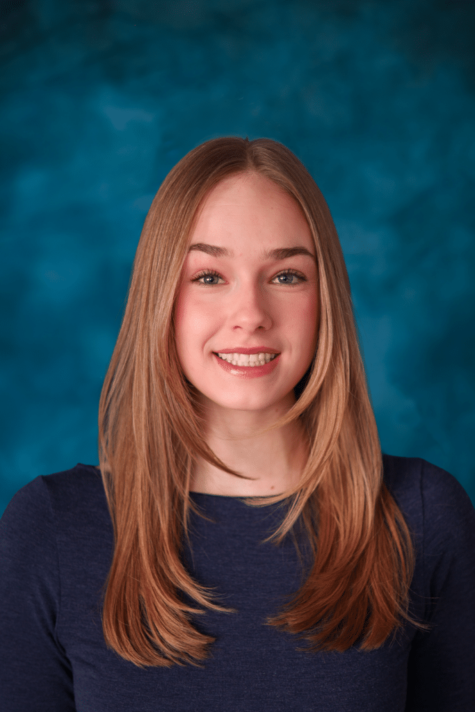 A young woman with straight blonde hair smiles in front of a blue background, wearing a navy top.