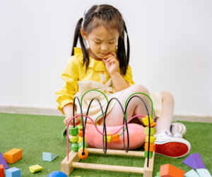 Young girl in yellow sits on grass, playing thoughtfully with a wooden bead maze and colorful blocks.