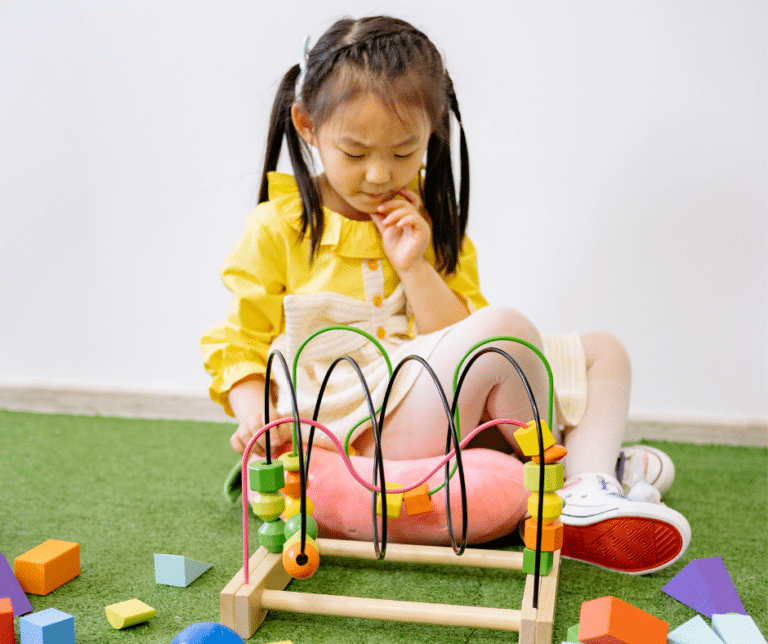 Young girl in yellow sits on grass, playing thoughtfully with a wooden bead maze and colorful blocks.