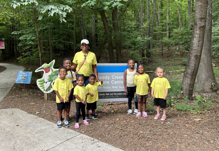 A group of kids and an adult pose by a "Reedy Creek Nature Center" sign in a wooded area.
