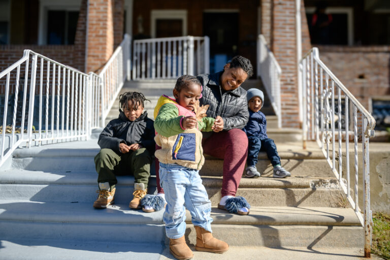 A woman and three children sit together on outdoor steps in front of a brick house on a sunny day.