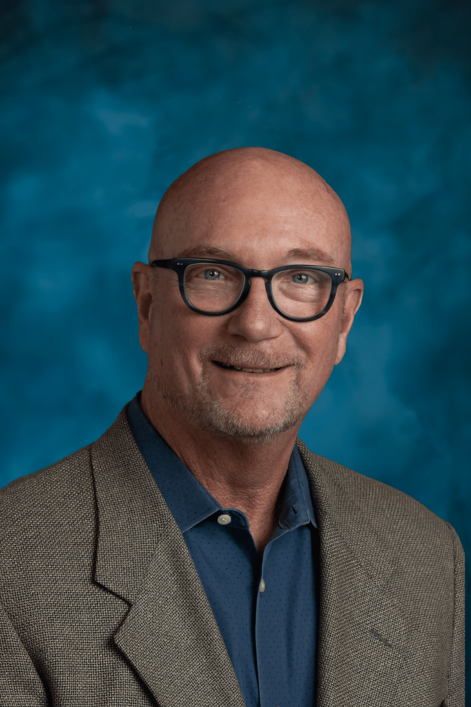 Smiling bald man with glasses in a blazer and blue shirt, in front of a blue mottled background.