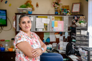 Woman in a floral shirt stands smiling with arms crossed in a colorful, cluttered home office space.