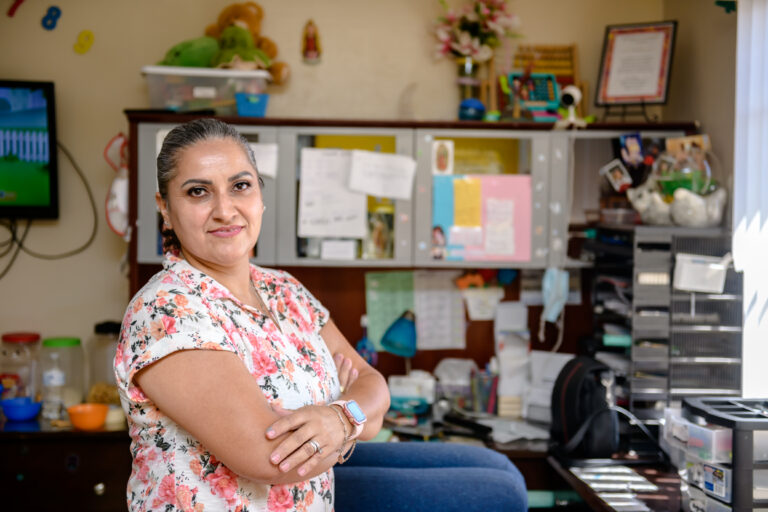 Woman in a floral shirt stands smiling with arms crossed in a colorful, cluttered home office space.