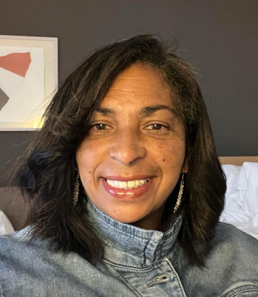 Woman smiling, wearing a denim jacket and earrings, with dark hair, indoors with art and a bed in the background.