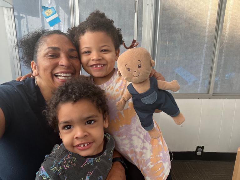 Smiling adult with two young children and a doll, posing for a cheerful selfie indoors.