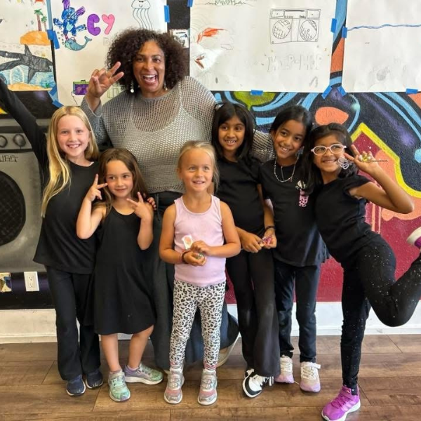 A smiling woman and six young girls pose together, making peace signs in front of colorful wall art.