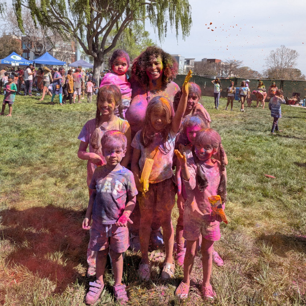 A group of kids and an adult, covered in colorful powder, celebrate Holi outdoors on a sunny day.