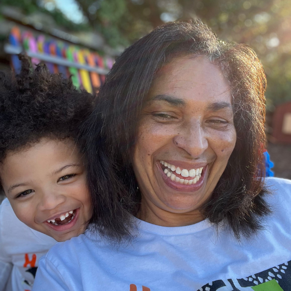 Smiling woman and child pose closely together outdoors in bright sunlight, both laughing happily.