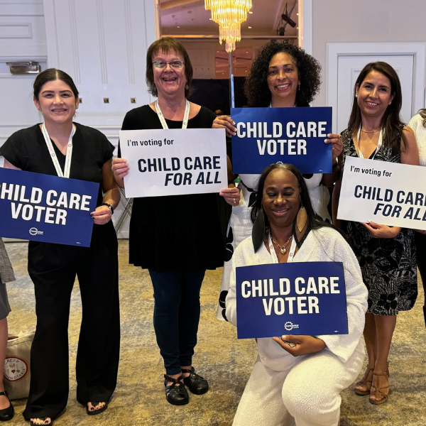 Five women smiling and holding signs supporting child care and voting for child care for all at an indoor event.