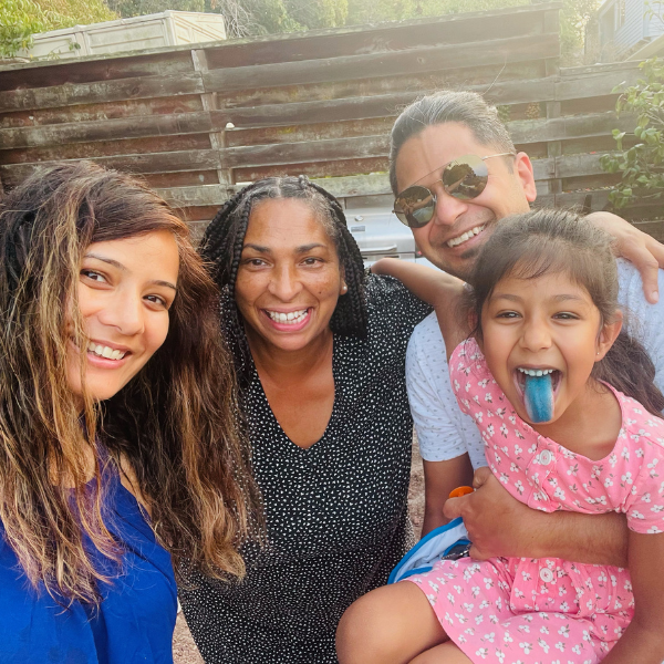 Four smiling people pose outside; a girl in a pink dress has a blue tongue and sits on a man's arm.